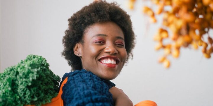 Happy young black woman holding basket with lettuce on shoulder and cluster of yellow dates in grocery store on gray background