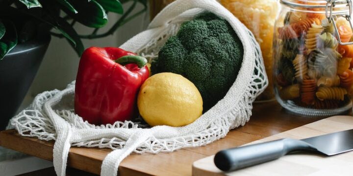 Composition of ripe fresh vegetables in net bag placed on kitchen counter near jars with pasta and chopping board with knife