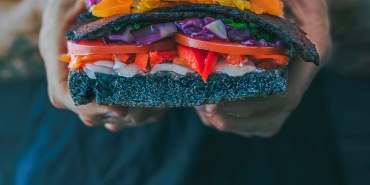 person holding meat sandwich with sliced tomatoes, onions, and grilled meat